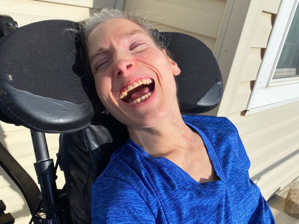 A woman in a wheelchair with gray hair wearing a blue shirt smiles widely.