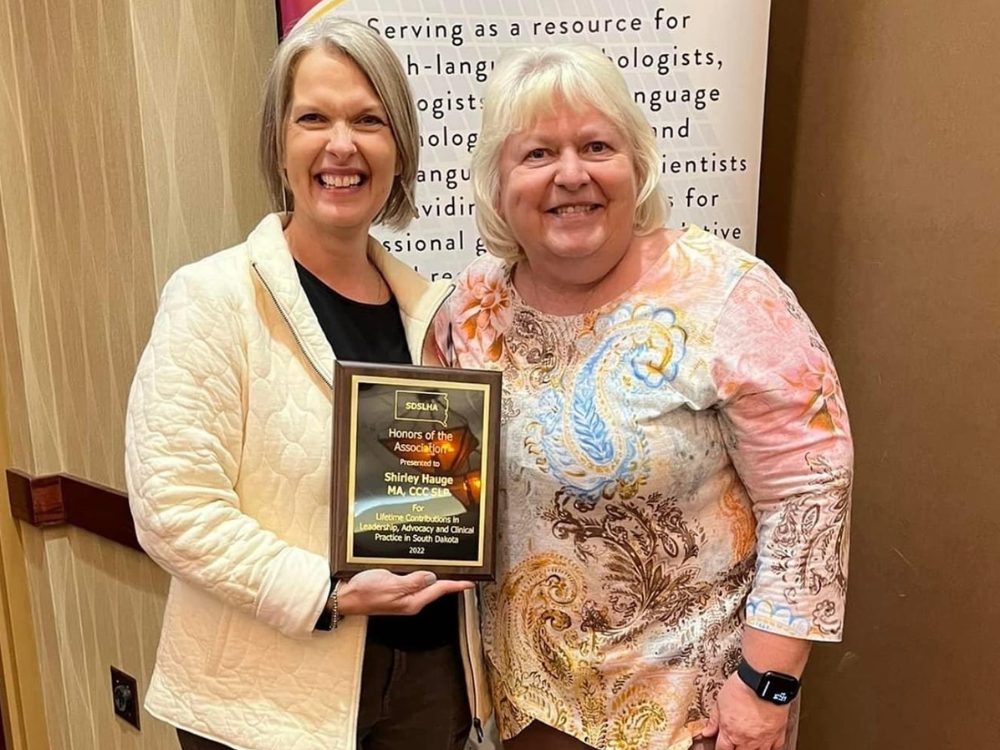 One woman with gray hair holding an award plaque stand next to a woman with white hair. Both women are smiling at the camera.