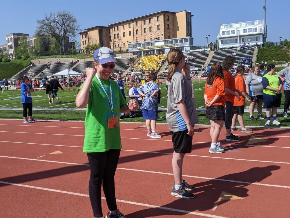 A woman wearing a hat and green shirt stands on a track with other participants getting ready for a race.