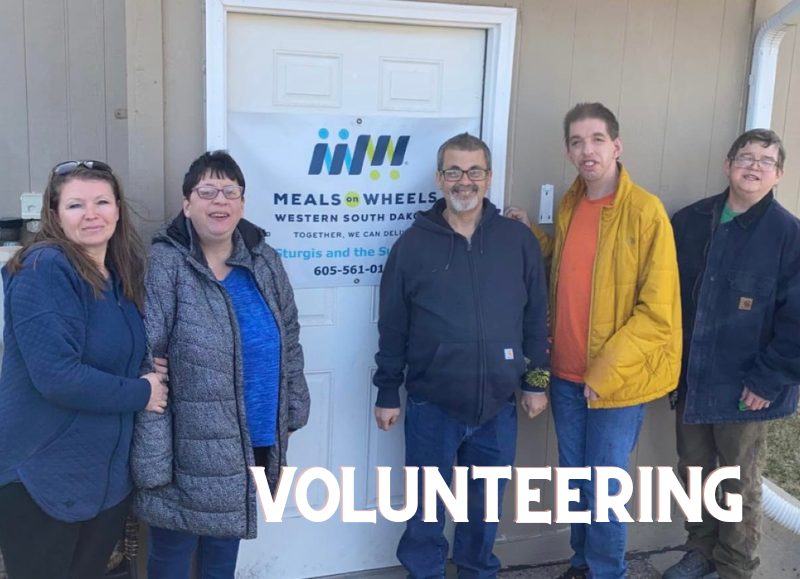 Three men and two women stand in front of the Meals on Wheels sign.