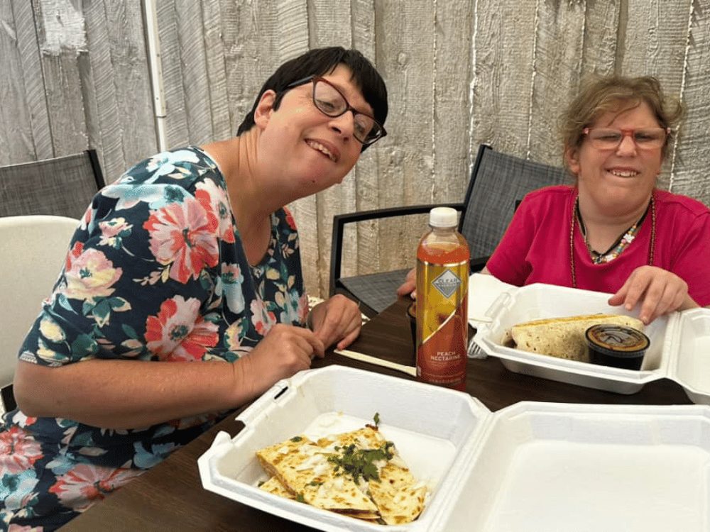 Two women with short brown hair and glasses are seated at a table. They are smiling. They both have takeaway containers with Mexican food from Mercy's Taco Truck.