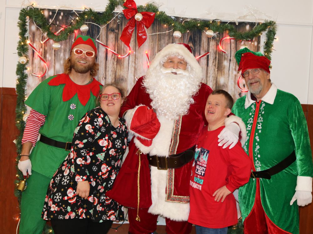 Four people dressed in green and red standing with their arms around each other with Santa Claus in the middle